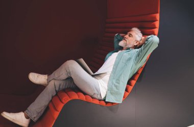 Üst düzey Kafkasyalı işadamı modern ofis salonunda dinleniyor, kucağında dizüstü bilgisayarla çalışmayı yansıtıyor, iş-yaşam dengesini ve rahat bir çalışma ortamında stratejik düşünmeyi sembolize ediyor