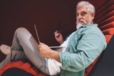 Tabletle rahat poz veren son sınıf erkek profesyonel, modern ofis mobilyalarının üzerine oturmuş, düşünceli, stratejik bir ifadeyle, bir iş ortamında teknoloji kullanımının rahatlığını ve kolaylığını temsil ediyor.