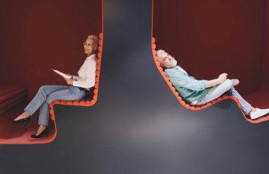 Günlük iş ortamında iki neşeli kıdemli profesyonel; belgeli kadın işkadını ve dizüstü bilgisayarlı erkek işadamı, yaratıcı ofis salonunda oturmuş, takım çalışması ve konfor tasvir ediyor.