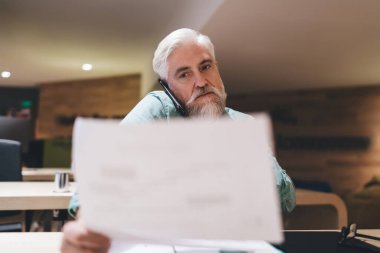Beyaz sakallı, deniz mavisi bir gömlek giyen, ofis ortamında odaklanma ve verimlilik gösteren, telefon ve belgelerle birden fazla iş yapan Kafkasyalı kıdemli işadamı.