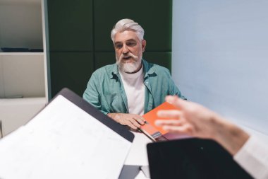 Beyaz sakallı, kıdemli, beyaz sakallı bir beyaz adam. İşbirlikçi ofis ortamında evrakları alıyor. Üzerinde mavi bir gömlek var ve düşünceli bir davranış sergiliyor.