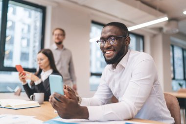 Beyaz gömlekli ve gözlüklü neşeli genç Afro-Amerikan erkek çalışanı iş günü iş yerinde iş arkadaşlarıyla mesajlaşırken kameraya gülümsüyor.
