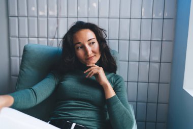 Genç, dalgın, siyah saçlı, elleri çenesinde beyaz fayanslı duvara yaslanmış rahat koltukta oturan ve düşünceli bakışlarını çeviren bir kadın.