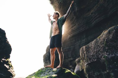 Alçak açılı, gündelik kıyafetli, şortlu genç bir erkek. Güneşli bir günde, kollarını kaldırıp, muzaffer bir kutlama yapmak için tepeden tırnağa kayalıklara bakıyor.