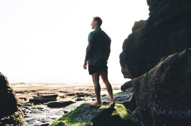 Erkek gezginin tüm vücudu yosun kaplı kayanın üzerinde duruyor ve sakin denizin engin genişliğine bakıyor.