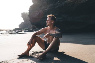 Siyah şortlu, kolları dövmeli genç turistin, güneşli bir günde tatilin tadını çıkarırken, kumlu sahilde dizleriyle oturduğu ve gözlerini kaçırdığı tüm vücut manzarası.