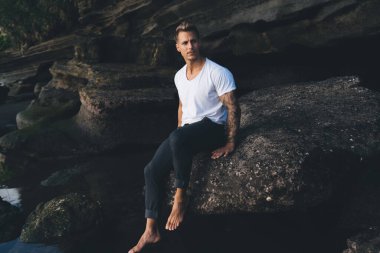 Ciddi sakallı, beyaz tişörtlü, kolları dövmeli genç bir erkek. Akşamları kameraya bakarken Strea 'nın üzerinde kayalık bir kayanın üzerinde oturuyor.