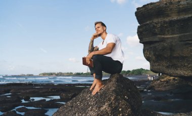 Düşünceli genç turistin tüm vücudu deniz kıyısında bir taşın üzerinde oturmuş kitap tutarken mavi gökyüzüne bakmıyor.