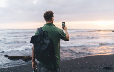 Sırt çantalı tanınmayan bir erkek gezginin dalgalı denizde akşamları kumlu sahilde dururken renkli günbatımının fotoğrafını çekerken görüntüsü.