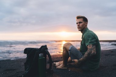 Spor kıyafetleri ve dövme koluyla kumlu sahilde çantalı ve su şişeli bir şekilde oturmuş dalgalı deniz ve günbatımı gökyüzüne bakan genç bir adam.