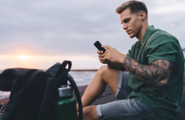 Günbatımında dinlenirken kıyıda oturan ve cep telefonunu karıştıran dövmeli genç bir erkeğin yan görüntüsü