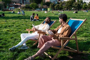 Gülümseyen genç, sakallı homoseksüel İspanyol erkek arkadaşları sohbet ederken ve yeşil çimenlikteki şezlonglarda dinlenirken ve ziyaretçilere ve ağaçlara karşı kitaplar okurken yan görünüm