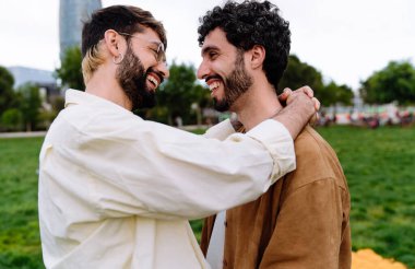 Kıvırcık saçlı, kollarıyla boynuna sarılan, birbirine bakıp gülen ve parkta kaynaşan neşeli sakallı eşcinsel çiftin yan görüntüsü.