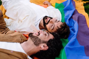 Gülümseyen genç, sakallı eşcinsel erkek LGBT bayrağında gözleri kapalı ve güneş ışığında yeşil parkta dinlenirken gözlerini kaçırıyor.