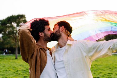 Mutlu genç eşcinsel çift, parkta yeşil çayırda dururken gökkuşağı LGBT bayrağını tutarken öpüşüp kucaklaşıyorlar.