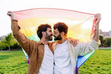 LGBT bayrağı tutan ve güneş ışığında yeşil çimlerde yan yana dururken öpüşen İspanyol, eşcinsel çifti sevmekten memnun oldum.