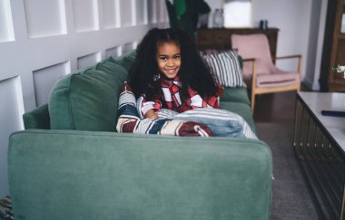 Keyifli Afro-Amerikan kıvırcık saçlı kız rahat oturma odasındaki yastıklarda dinlenirken gülümseyen ve kameraya bakan.