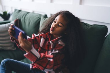 Yüksek açılı, kıvırcık saçlı, kanepede oturan ve akıllı telefondan selfie çekerken sivri ağızlı Afro-Amerikan kadın çocuk.