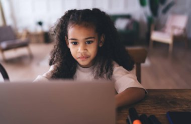 Evdeki iş saatinde ekranlara bakarken tahta masada dizüstü bilgisayarla oturan, günlük giysili, kıvırcık saçlı, tatlı Afrikalı küçük bir çocuk.