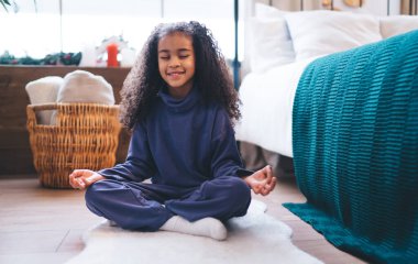 Huzurlu bir evde meditasyon yapan Afro-Amerikalı genç bir kız gözleri kapalı bir şekilde yan yana oturuyor ve nazikçe gülümsüyor, farkındalığı ve iç huzuru teşvik ediyor..