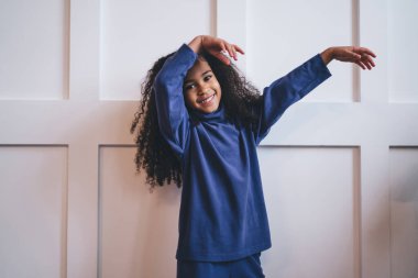 Neşeli Afro-Amerikalı kız mavi gece elbisesi giyip dans ederken modern odadaki beyaz arka planda kameraya bakarak dans ediyor.