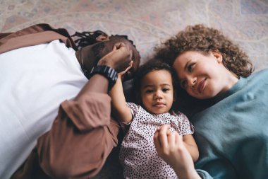 Modern oturma odasında birlikte vakit geçirirken ve birbirlerine bakarken gündelik giysiler içinde sırt üstü yatıp el ele tutuşan çok ırklı ailelerin yüksek açısı.