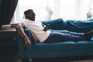 Afro-Amerikan sakallı, günlük kıyafetlerle kanepeye oturmuş, akıllı telefonları karıştırıyor ve evdeki rahat, parlak oturma odasında dinleniyor.