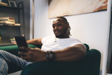 Pozitif sakallı ciddi Afro-Amerikan erkek, gündelik giysiler içinde kanepede oturuyor ve rahat bir odada duvarları ve modern apartmandaki rafları karıştırırken ekrana bakıyor.