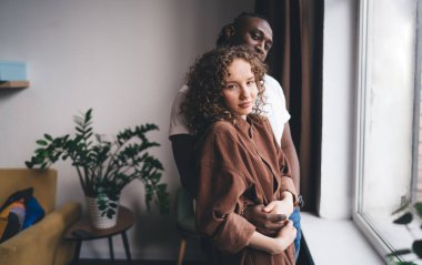 Romantik, çok kültürlü çiftler, gündelik giysiler içinde, pencerenin kenarında saksı bitkileriyle dikilip kameraya bakarken romantik bir şekilde sarılıyorlar ve oturma odasında birlikte vakit geçiriyorlar.