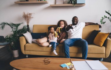 Mutlu, genç, çok ırklı ebeveynler ve günlük kıyafetleri içinde tatlı bir kız çocuk. Kanepede uzaktan kumandayla oturup televizyon izlerken gülümseyip muz yiyorlar.