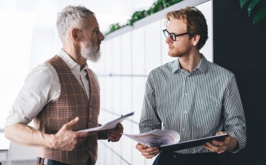 Odaklanmış erkek iş ortakları ellerinde belgeler ve kırpıcıyla ofiste duruyor ve parlak güneş ışığıyla dolu bir odada proje çalışmaları hakkında tartışırken birbirlerine bakıyorlar.