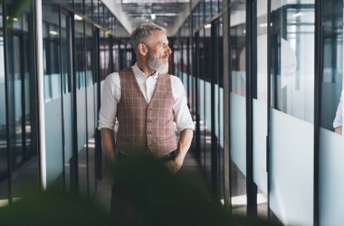 Kendine güvenen, gri saçlı, resmi kıyafetli bir işadamı elleri ceplerinde modern ofis merkezinin cam kapılarının yanında dikilirken yüzünü çeviriyor.