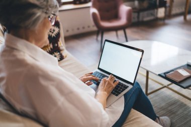 Gündelik giysiler ve gözlüklerle kanepede oturan, şirket tarafından dizüstü bilgisayarla gönderilen belgeleri inceleyen ve bulanık oturma odasındaki ekrana bakan kimliği belirsiz yaşlı kadın serbest yazar.