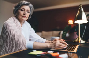 Masada dizüstü bilgisayarla oturan ve uzaktan proje üzerinde çalışırken aşağıya bakan serbest çalışan olgun bir kadın.