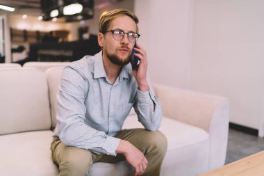 Modern açık uzay ofisindeki koltukta oturmuş cep telefonuyla konuşan şık günlük kıyafetler ve gözlüklerle genç, sakallı bir girişimci.