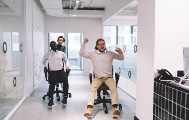 Çağdaş çalışma alanında birlikte eğlenirken, çeşitli meslektaşlarıyla sandalyeleri yuvarlayan tam vücut kadın ofis çalışanları.
