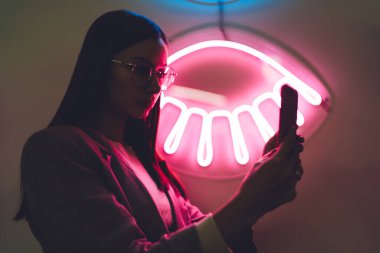 Gözlüklü ve ceketli genç bir kadın, pembe neon ışığın yanında dururken cep telefonu kullanarak internette geziniyor.