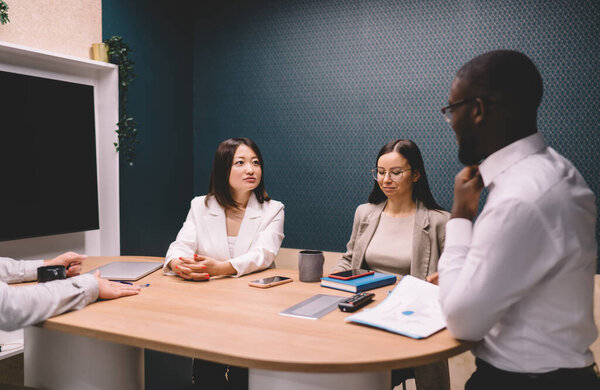 Группа мультиэтнических молодых коллег в формальных костюмах собирается за столом и обсуждает детали проекта во время совместной встречи во время деловой конференции