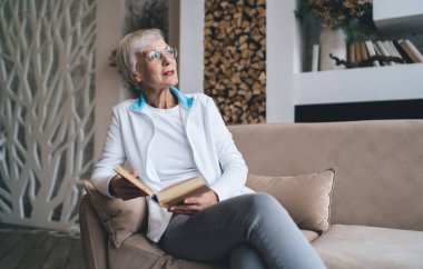 Düşünceli, gözlüklü, kanepede dinlenen ve boş zamanlarında ilginç kitaplar okuyan, evdeki rahat oturma odasına bakan son sınıf öğrencisi bir kadın.