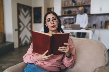 Rahat bir dairede dinlenirken, kitap okuyup kitap okuyan, gündelik giyimli ve gözlüklü son sınıf öğrencisi bir kadın.
