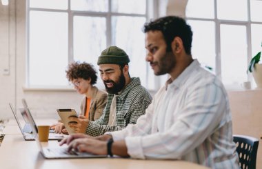 Çeşitli erkek ve kadın iş arkadaşlarının gündelik kıyafetleriyle masadaki dizüstü bilgisayarları ve tabletleriyle modern çalışma alanındaki projeleri incelerken ve çalışırken yan görünümü
