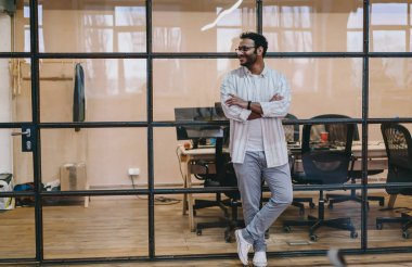 Moda kıyafetleri ve gözlükleriyle, kolları ve bacakları çaprazlanmış cam duvara yaslanmış, yaratıcı çalışma alanında dinlenen genç sakallı bir erkek vücudu.