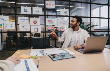 Pozitif sakallı İspanyol erkek işçi gündelik giysiler içinde masada dizüstü bilgisayarla ve tabletle oturmuş el kaldırarak konuşuyor ve modern ofiste çalışıyor.