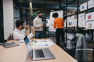 Ofisteki projenin detaylarını analiz ederken ve tartışırken, çizelgelerle ve şemalarla yan yana duran sıradan giyinen erkek arkadaşlarıyla yüzsüz bir kadının arka plan görüntüsü.
