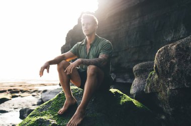 Güneşli bir günde kayalık kıyılarda kayalıklarda dinlenirken kayaların üzerinde oturan ve başka yöne bakan kendinden emin genç bir erkek.