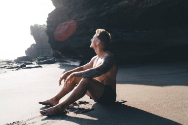 Çıplak ayaklı, şortlu genç bir erkeğin güneşli bir günde gölgesiyle kayalık sahildeki kumlu kumsalda oturuşu.