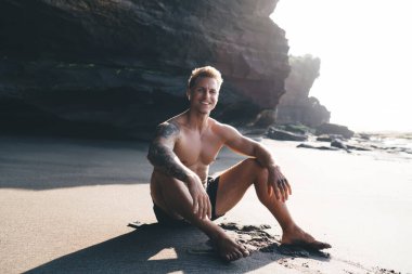 Siyah şortlu ve kollu genç bir erkeğin kumlu sahilde oturup yaz tatilinin tadını çıkarırken kameraya bakışıyla.