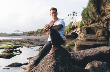 Nehrin kayalık kıyılarında güneşli bir günde kameraya bakarken, günlük giysiler içinde kitap tutan sakallı bir erkek.