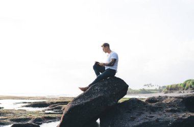 Kitap okurken ve güneşli bir günde tatilin tadını çıkarırken, nehir kenarındaki kayalık arazide, taşa yaslanmış gündelik giysiler içinde genç bir erkeğin alçak açılı yan görüntüsü.