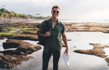 Spor ayakkabıları ve güneş gözlükleriyle kumlu sahilde el sallayan ve kameraya bakan pozitif erkek gezgin.